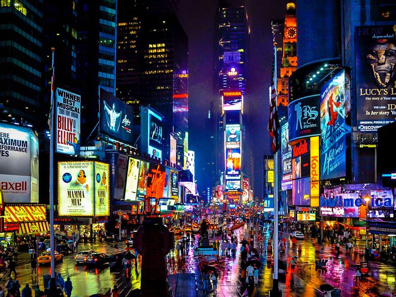 Times Square at night, illuminated by bright lights and billboards, with a wet street reflecting the colorful scene.