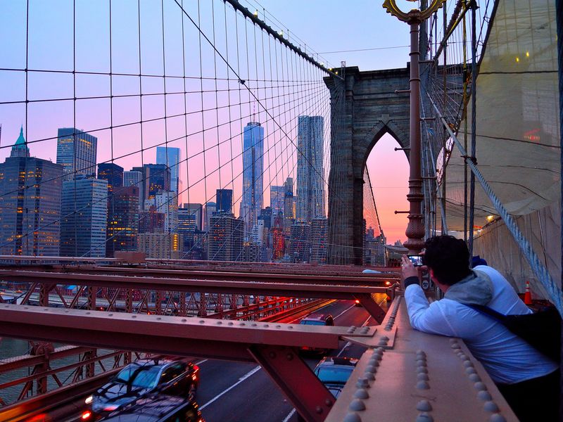 A person taking a photo of the New York City skyline from the Brooklyn Bridge at sunset.