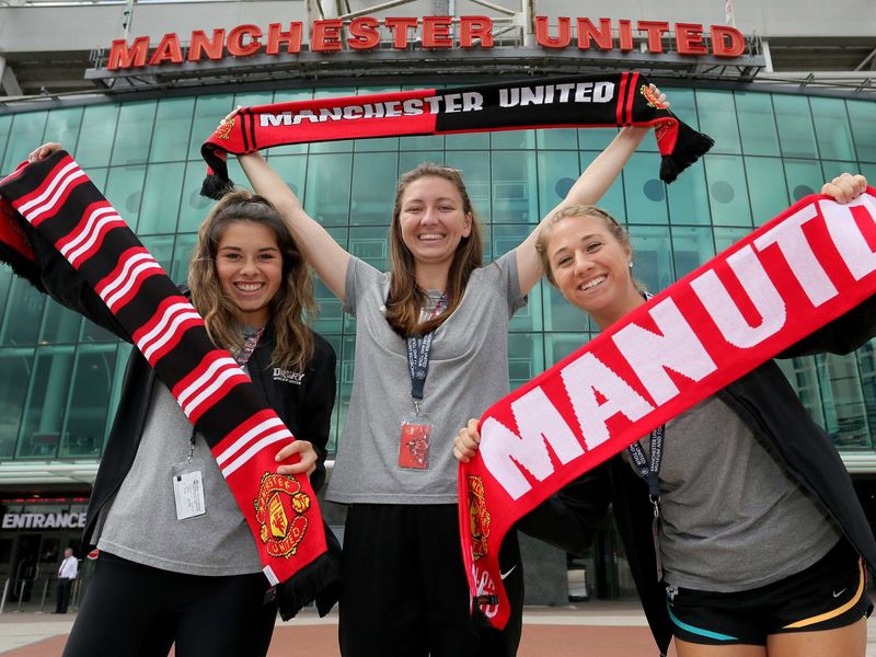 Three excited young adult women proudly display their Manchester United scarves outside Old Trafford stadium.