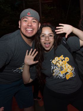 Three young adults playfully pose for a photo, their smiles and energetic hand gestures conveying a sense of shared enjoyment. Their matching gray t-shirts suggest they are part of a group or event.