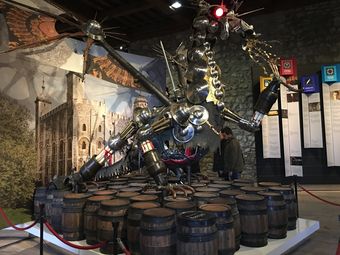 Large metal dragon sculpture in a museum, with a castle mural in the background.