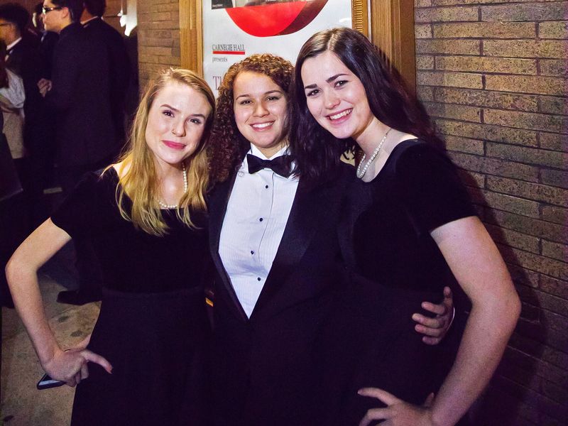 Three young adults dressed in formal attire stand below the Carnegie Hall sign.